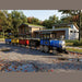 Frontier Trackless Train   (blue, yellow, red, black) parked outdoors on a paved road in front of a modern building with a green roof.