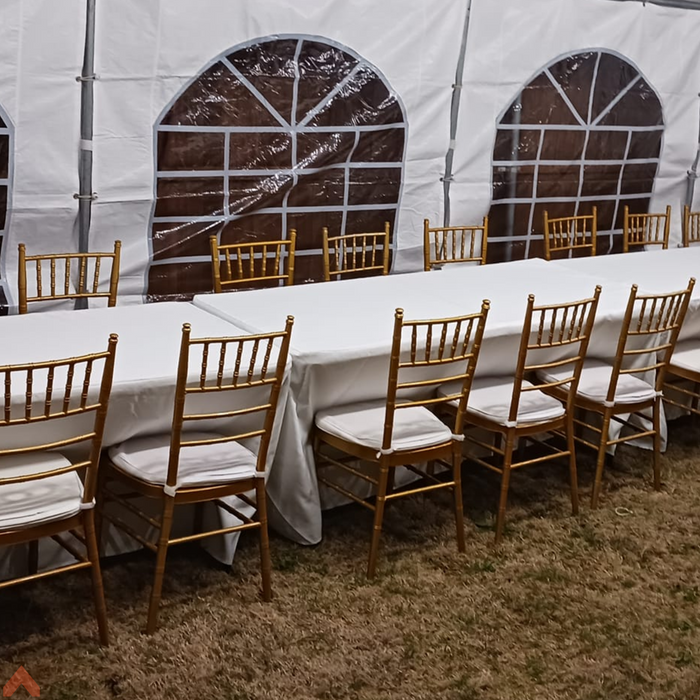 Indoor event setup inside a white frame tent with cathedral window sidewalls, featuring gold resin Chiavari chairs with white seat cushions lined up along long tables covered in white tablecloths