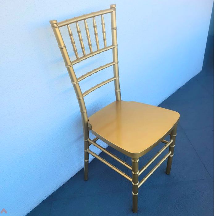 Beyond Tent Hercules Premium Resin Chiavari Chair. Three-quarter front view of a gold resin Chiavari chair featuring a bamboo-style spindle backrest and a smooth solid seat, displayed on a blue surface against a light blue backdrop