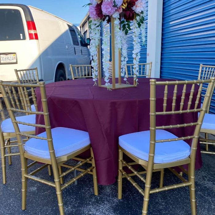 Outdoor event setup featuring gold resin Chiavari chairs with white seat cushions arranged around a round table covered in a burgundy tablecloth, topped with a tall floral centerpiece