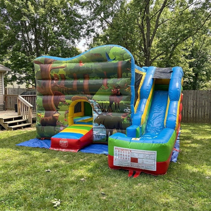 A large inflatable children's bounce house and slide set set up on a green grass backyard. The structure features a forest-themed front wall with moose graphics, a rainbow-striped entry ramp, and a blue slide with yellow side accents.