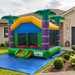 Beyond Tent Lightweight Palm Tree Bouncer, purple and green castle bounce house with palm tree towers on all four corners, set up in a front yard next to a house