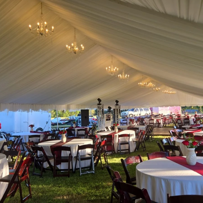 Large frame tent interior with white pleated ceiling liner, a row of crystal chandeliers along the ridge, round tables with white linens and burgundy runners, and dark wood folding chairs with red cushions
