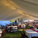 Large frame tent interior with white pleated ceiling liner, a row of crystal chandeliers along the ridge, round tables with white linens and burgundy runners, and dark wood folding chairs with red cushions
