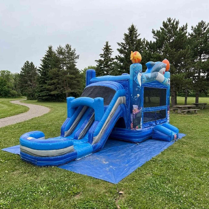 Ocean Shark Bounce House Combo  set up on a grassy lawn outdoors with trees and a cloudy sky in the background. Green grass grey sky. 