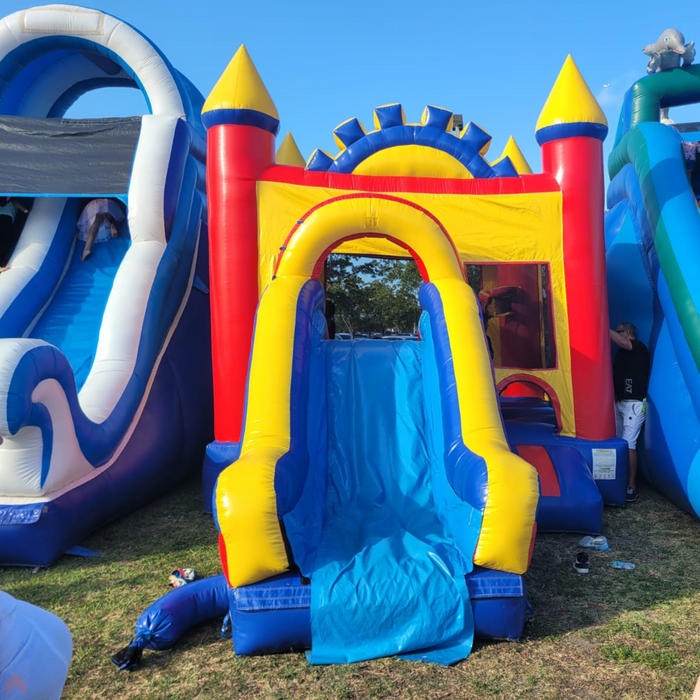 A professional setup of the Rainbow Play Wet & Dry inflatable combo on grass, positioned alongside other rental units. View highlights the vibrant blue slide, red castle walls, and the compact footprint of the commercial-grade bounce house.