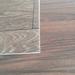 Close-up angled view of a Rustic Oak vinyl flooring sample's edge laid against a reddish wood-grain surface, showing the plank thickness and texture detail