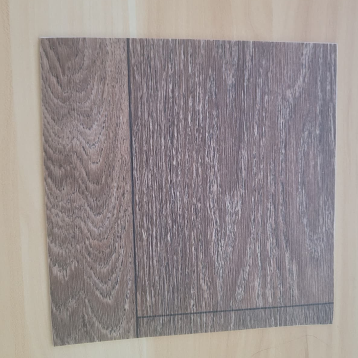Rustic Oak vinyl flooring plank sample laid diagonally on a light natural wood surface, showing the brown wood-look grain pattern from an angle