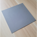 A square gray textured tile or mat shown at an angle on a light wood grain surface.