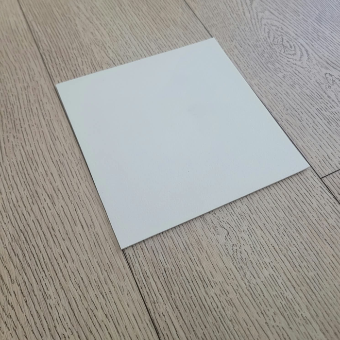 Matte White Rolled Vinyl Flooring sample swatch placed diagonally on a light oak wood plank floor, angled perspective view