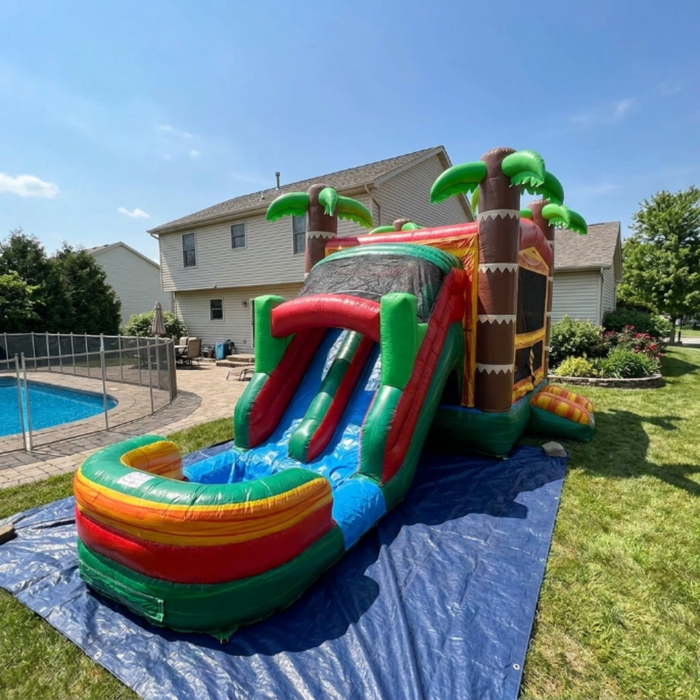 Tiki Trail Dual Lane Wet and Dry Combo inflated on a blue tarp in a bright sunny backyard. Full unit from front-left — dual water slide lanes and multi-color splash pool on the left, bounce house with large mesh viewing window, yellow-orange walls, and tiki-column corners on the right, three green palm trees on top. A fenced in-ground swimming pool, large house with light siding, and blue summer sky with clouds visible in the background.