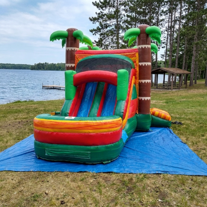 Tiki Trail Dual Lane Wet and Dry Combo on a blue tarp at a scenic lakeside setting with green and yellow grass. Front-left view — dual water slide section and colorful splash pool on the left, bounce house with mesh window and brown tiki column corners on the right, three green palm trees at the top. A wooden lake pavilion and pine trees line the shoreline behind the unit. Partly cloudy sky. Serene outdoor rental setting.