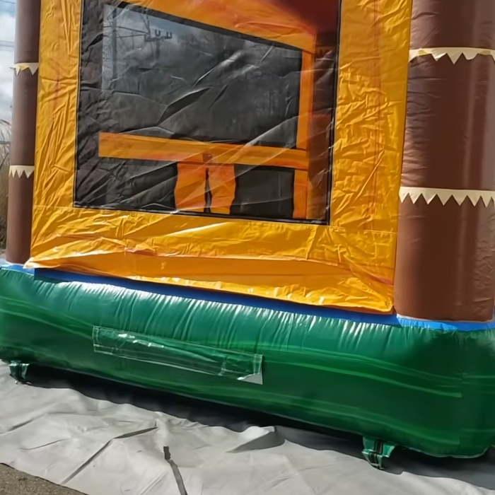 Close-up interior entrance of the Tiki Trail Dual Lane Wet and Dry Combo bounce house, shot from floor level looking into the jump area. Shows yellow and orange inflatable walls with brown tiki column side posts and white sawtooth-pattern decorative trim at the entry threshold. Dark mesh window panel visible inside. Photographed indoors on white tarp, highlighting the commercial PVC seam quality and reinforced entry passage construction.