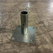 A galvanized steel mounting bracket consisting of a square plate with four pre-drilled holes and a vertical cylindrical pipe section, shown on a scratched concrete floor.