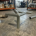 A galvanized steel T-shaped angle bracket support leg with a vertical post and two horizontal arms, shown on a concrete workshop floor with metal pipes and storage shelves in the background.