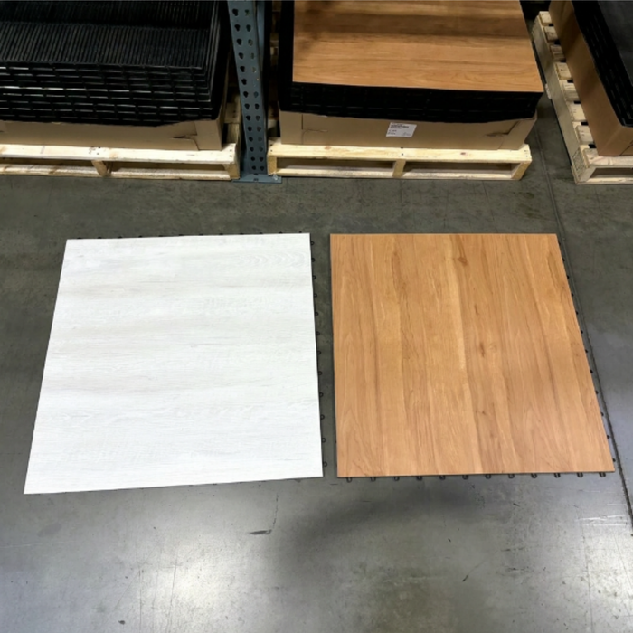 Two square flooring samples displayed side-by-side on a warehouse floor, one with a white textured finish and the other with a light brown wood grain finish. This is a Beyond Tent exclusive picture to show the real colors of the product. 