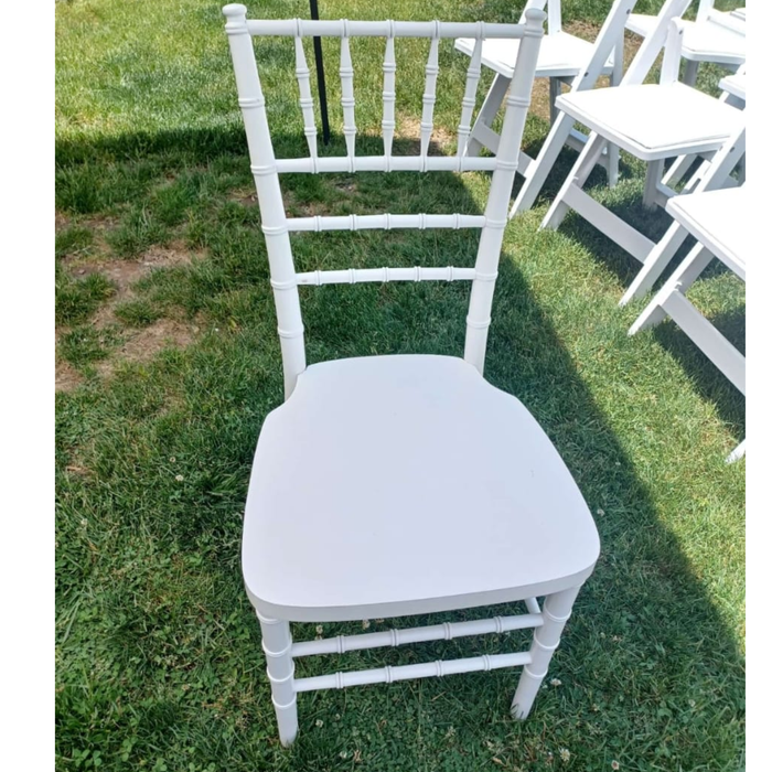 "Front view of a white resin Chiavari chair with a spindle backrest and flat seat, set outdoors on a green grass lawn alongside other event chairs."