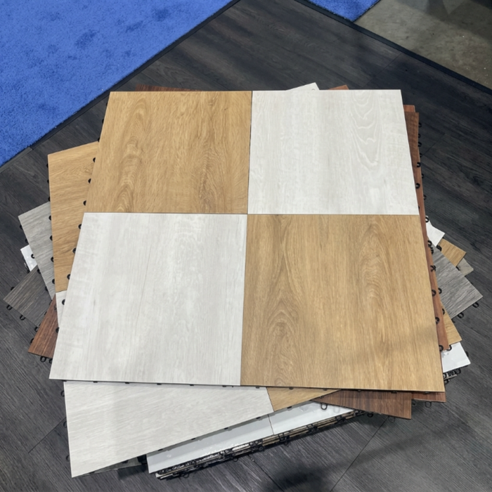 A stack of square interlocking floor tiles or flooring samples shown from above on a dark wood floor, with some blue flooring samples visible in the background. The top sample is split into four quadrants: two light natural wood-grain panels and two off-white/light beige panels. Beyond Tent Picture. 