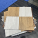 A stack of square interlocking floor tiles or flooring samples shown from above on a dark wood floor, with some blue flooring samples visible in the background. The top sample is split into four quadrants: two light natural wood-grain panels and two off-white/light beige panels. Beyond Tent Picture. 