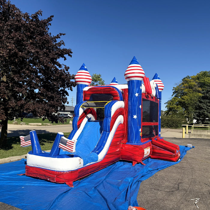 American Titan Wet and Dry Combo set up outdoors on a blue tarp, blue slide with white rails into a splash pool with two American flags, red marble vinyl and striped dome towers