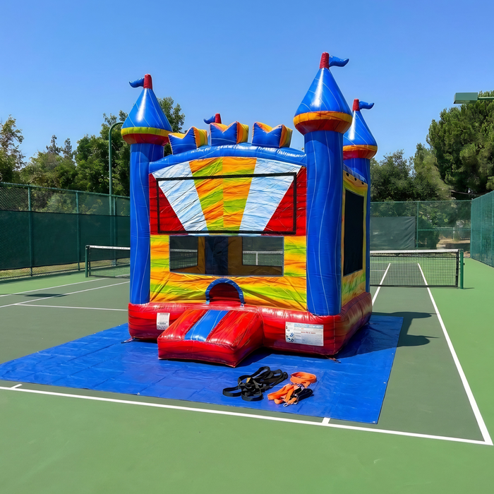 Front angle of the Beyond Tent 13x13 Wonder Castle commercial bounce house on a green tennis court, showing the blue safety tarp and a set of heavy-duty orange ratchet straps and black tie-down stakes in the foreground.