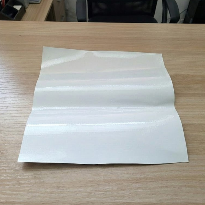 Close-up image of a white 16 oz coated blockout PVC vinyl fabric swatch laid flat on a light wood surface, illustrating the smooth texture and opaque quality of the material used for tent tops