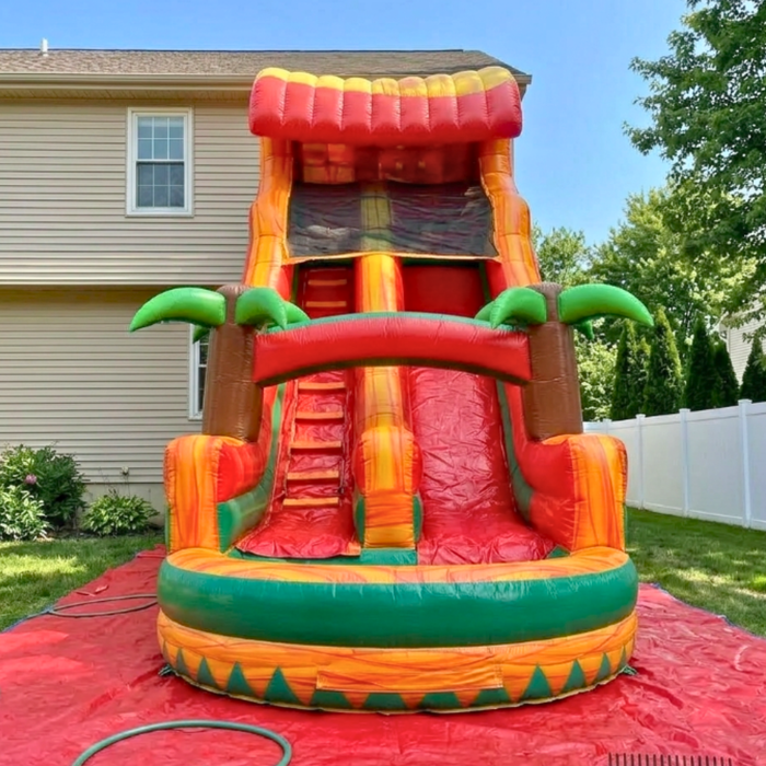 Alt Text: Beyond Tent 18-foot commercial Tropical Inflatable Water Slide with 3D palm trees and a wide landing pool, set up on a red protective ground tarp in a residential backyard with a white vinyl fence.
