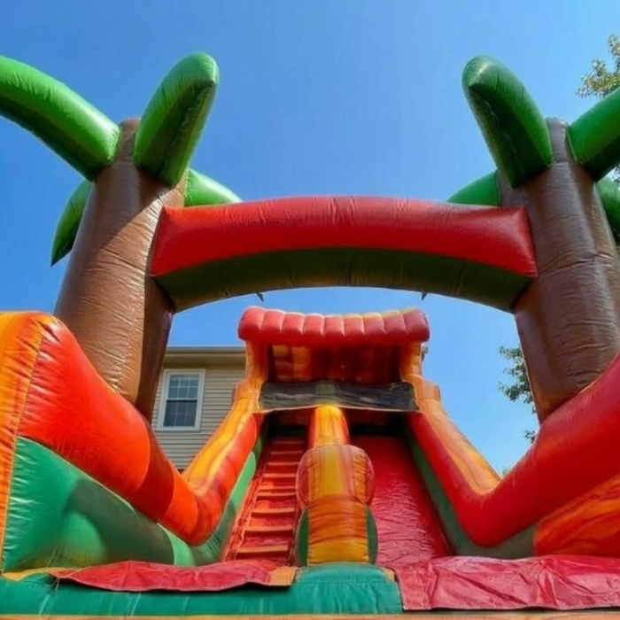 Low-angle view of the Beyond Tent 18-foot Tropical Water Slide showing the red inflatable climbing stairs, 3D palm tree pillars, and high-walled slide chute against a clear blue sky.