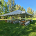Ground-level view of a Beyond Tent 30x40 Classic Series West Coast frame tent set up for an outdoor event, featuring rustic wooden farm tables and white folding chairs on a lush green lawn with a scenic forest background.