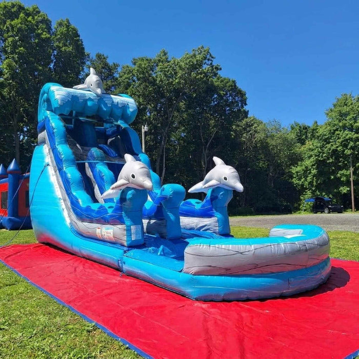 A professional installation of Beyond Tent's commercial-grade blue and white dolphin-themed inflatable water slide, featuring dual slide lanes, a safety splash pool, and 3D dolphins, set up on a red protective ground tarp in a residential backyard with lush green trees.