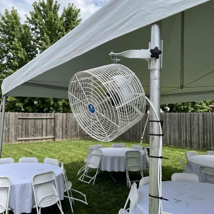 A large white outdoor tent fan mounted on a white tent pole in a backyard setup with white tables and chairs under a canopy.