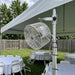 A large white outdoor tent fan mounted on a white tent pole in a backyard setup with white tables and chairs under a canopy.