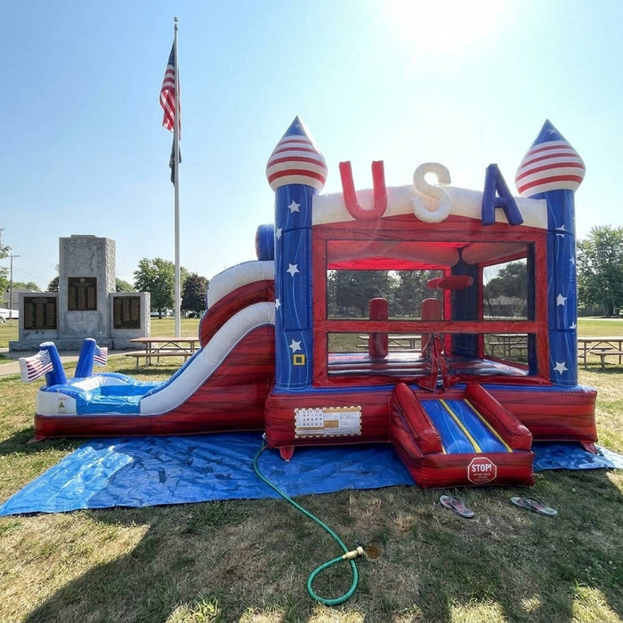 American Titan Wet and Dry Combo set up on a blue tarp in a grassy park with a flagpole, front entrance ramp, USA letters, mesh bounce windows, slide and splash pool connected to a hose