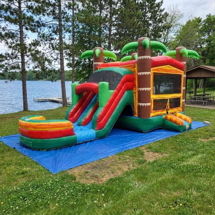 Tiki Trail Dual Lane Wet and Dry Combo fully set up at a lakeside park on a blue tarp over green grass. Full 29-foot unit visible from the front — dual water slide lanes with red and green stripes on the left, large bounce house with mesh window panel in the center, three large green palm trees on top. A scenic lake, tall pine trees, wooden dock, and covered picnic pavilion are visible in the background under a cloudy sky.