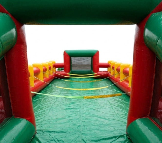 Interior view of a commercial inflatable human foosball arena showing the green vinyl playing floor, red side walls, yellow safety ropes, and a built-in soccer goal net.