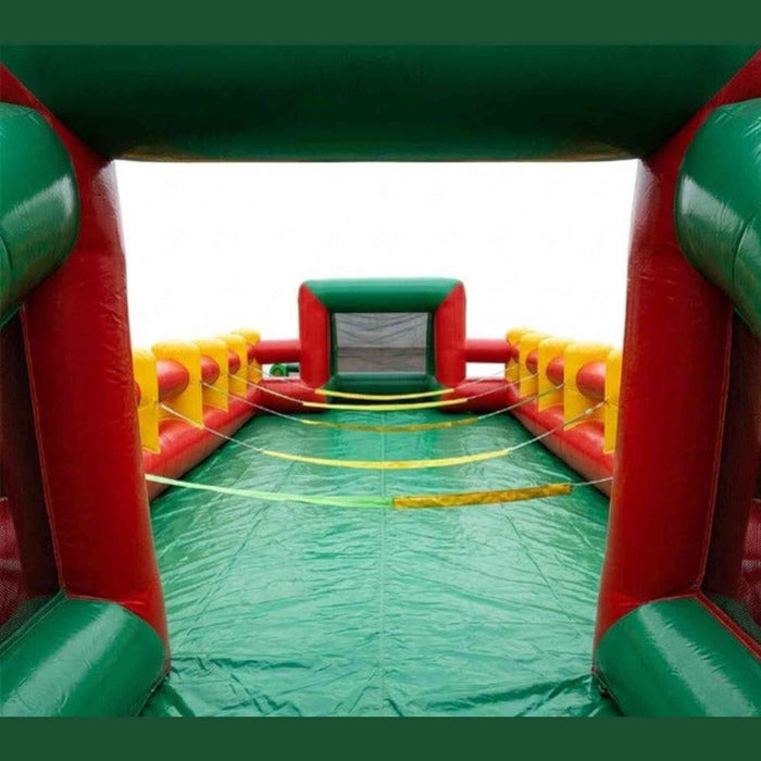 Interior view of a commercial inflatable human foosball arena showing the green vinyl playing floor, red side walls, yellow safety ropes, and a built-in soccer goal net.