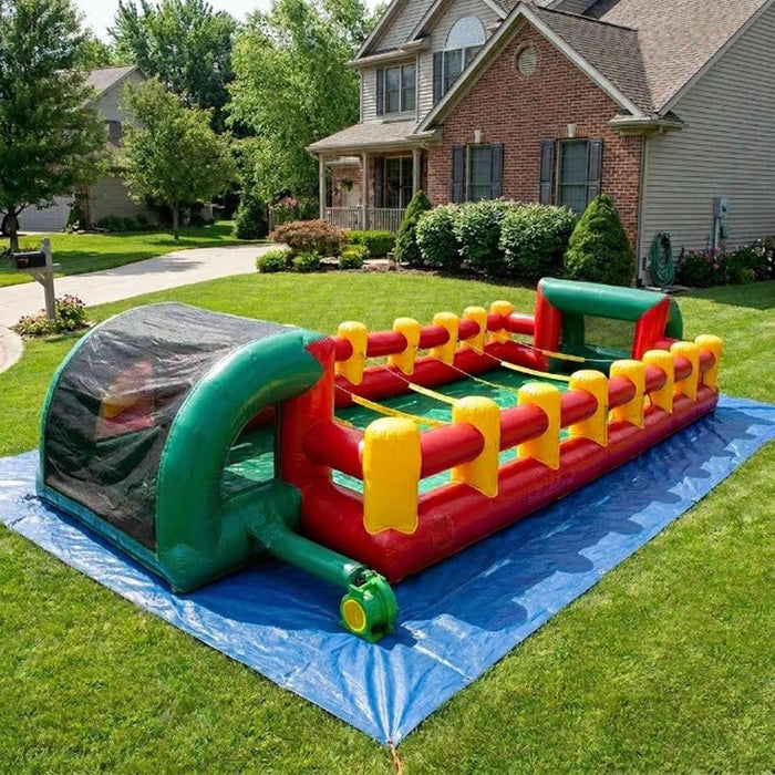 Commercial-grade inflatable human foosball arena set up outdoors on a blue tarp over a grassy lawn, showing its full length, green and red vinyl side walls, and yellow pole-connector straps.